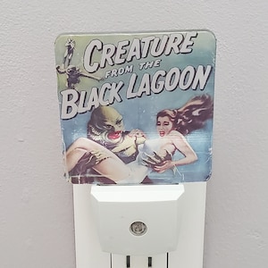 May include: A white electrical socket with a nightlight plugged in. The nightlight features a vintage movie poster design with the text "Creature from the Black Lagoon".