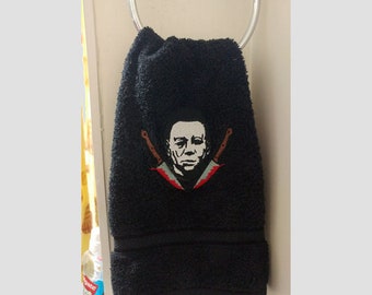 May include: Black hand towel with an embroidered image of Michael Myers from the Halloween franchise. The image includes two crossed knives with red accents.