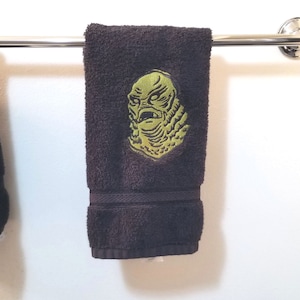 May include: A dark brown hand towel with a green embroidered image of the Creature from the Black Lagoon.