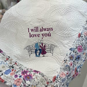 以下が含まれることがあります： 花柄の縁取りが施された白いキルトの毛布。毛布には、ベンチに座っているカップルと「I will always love you」という文字が刺繍されています。