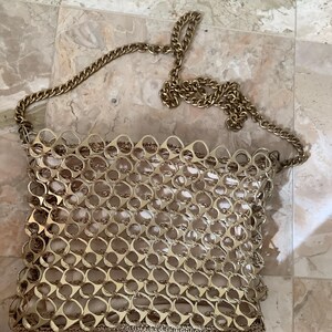 May include: A gold-colored handbag with a chain strap. The bag is constructed from interlocking metal rings, creating a unique, textured surface. The chain strap is also gold-toned, complementing the bag's overall aesthetic. The bag is likely a fashion accessory.