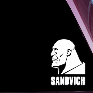 May include: White vinyl decal featuring a stern, bald man's profile with the word "SANDVICH" in bold letters below. The decal is likely for a car window or similar surface.