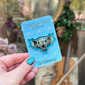 May include: An acrylic pin with a monster design, featuring a snarling face with blue and green eyes, and bat-like wings. The pin is on a light blue card with the text "Haunting Dance Acrylic Pin" and "KillVannah.com".