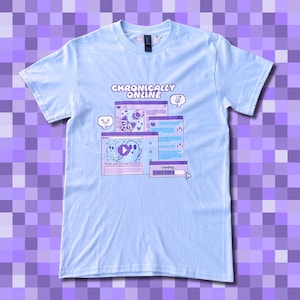 Peut inclure: T-shirt bleu clair avec l'inscription "Chronically Online" au-dessus d'un graphisme de fenêtres d'ordinateur. Les fenêtres présentent des fantômes de dessins animés, des cœurs et des bulles de discussion en nuances de violet et de blanc. Le t-shirt a un col rond.