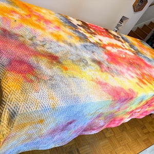 May include: A colorful, tie-dye patterned blanket or tablecloth. The textile features a woven texture and a vibrant mix of colors, including yellow, orange, pink, blue, and gray. The fabric is draped over a table.