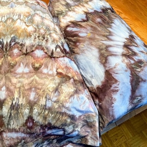 May include: Two tie-dye fabric pieces in shades of brown, beige, and blue. The fabric has a textured appearance with a unique pattern. The items are folded and lying on a wooden surface.