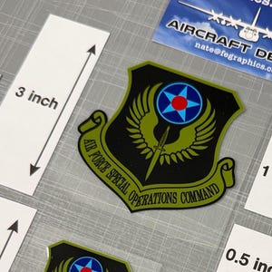 May include: A collection of Air Force Special Operations Command decals and stickers. The largest decal measures 3 inches in height. The image includes business cards with the text "Aircraft Decals" and the website "fegraphics.com".