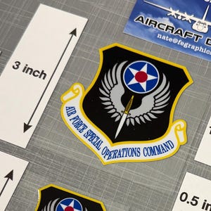 May include: Various sizes of Air Force Special Operations Command aircraft decals. The decals feature a black, yellow, and blue emblem with a star and wings design. Sizes range from 0.5 inches to 3 inches. The image also includes business cards.