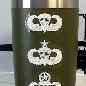 May include: A green YETI tumbler with three white parachute jump wings decals. The decals feature a star and a parachute. The tumbler is sitting on a table with a business card and a small American flag.