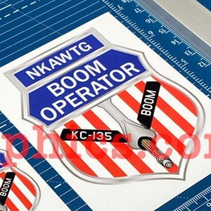 May include: Two stickers with a blue, red, and white shield design. The shield has the text "NKAWTG BOOM OPERATOR" in white letters on a blue background. The shield also has the text "KC-135" and "BOOM" in white letters on a red and white striped background.