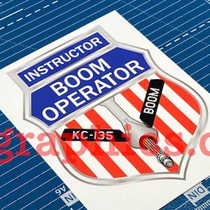 May include: A white sticker with a red, white, and blue shield design. The shield has the text "BOOM OPERATOR" in white letters on a blue background. The text "INSTRUCTOR" is above the shield. The text "KC-135" is on the shield. The text "BOOM" is on the right side of the shield.