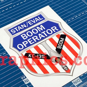 May include: A red, white, and blue shield-shaped sticker with the text "STAN/EVAL BOOM OPERATOR" in blue letters. The shield has a silver outline and a red, white, and blue striped background. The text "KC-135" is in a white rectangle on the shield. The text "BOOM" is in a black rectangle on the shield. The shield has a silver outline and a red, white, and blue striped background.