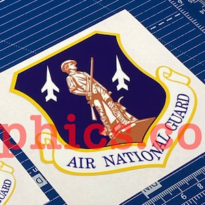 May include: Two white stickers with a blue and gold shield design. The shield features a soldier standing on a hill with two airplanes flying overhead. The text "AIR NATIONAL GUARD" is printed on the stickers.
