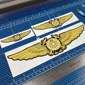 May include: Three gold-colored paper cutouts of pilot wings with a compass design. The wings are on a blue cutting mat with a ruler and a utility knife.
