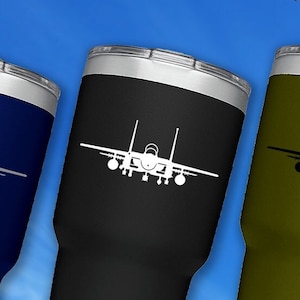 May include: Three stainless steel tumblers with a white silhouette of an F-15 fighter jet on each. The tumblers are in navy blue, black, and olive green. The text "Veteran Owned Business" is in the upper right corner of the image. The text "CUP NOT INCLUDED" is at the bottom of the image.