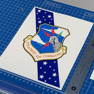 May include: A blue and white decal with a stylized lightning bolt and a fist holding a branch. The decal reads "Strategic Air Command".