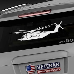 May include: White vinyl decal of a helicopter on a vehicle's rear window. The decal features the text "www.fegraphics.com". A "Veteran Owned Business" sticker with an American flag is also visible.