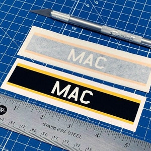 May include: Two black and yellow vinyl stickers with the word "MAC" printed in white letters. The stickers are on a blue cutting mat with a ruler and a craft knife.