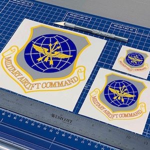 May include: Three white stickers with the words "Military Airlift Command" in yellow lettering. The stickers feature a blue globe with a yellow winged arrow design. The largest sticker is 7 inches wide and the smallest sticker is 3 inches wide.