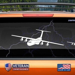 May include: White vinyl decal of a C-130 Hercules aircraft with lightning bolts on a car window. The decal is made in the USA and is a popular gift for aviation enthusiasts. "VETERAN OWNED BUSINESS" and "www.fegraphics.com Flight Engineered Graphics Aircraft Decals" are printed on the image.