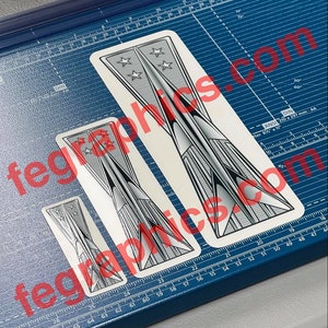 May include: Three stickers with a retro-futuristic design featuring a silver and black color scheme. Each sticker has a pointed shape with star accents and the text "fegraphies.com" in red. The stickers are on a blue cutting mat.