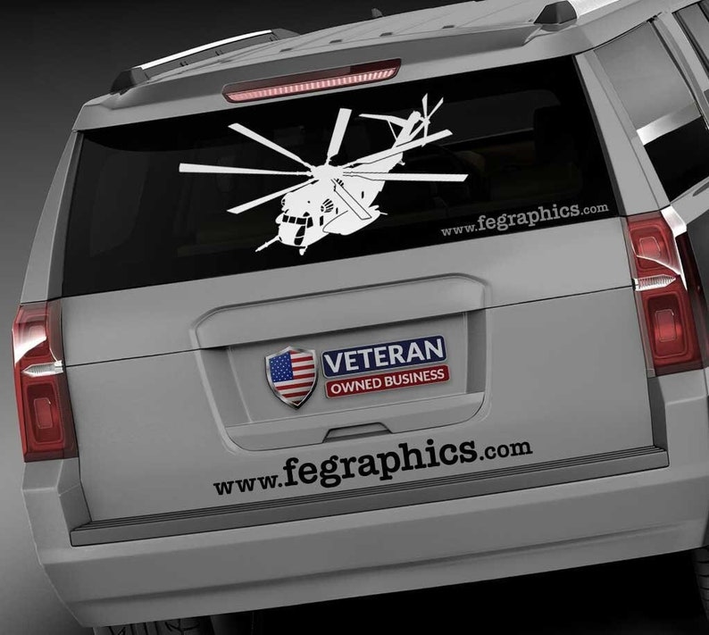 以下が含まれることがあります： グレーの車の後ろに、「Veteran Owned Business」のステッカーと「www.fegraphics.com」のウェブサイトが貼られた、白いヘリコプターのシルエット。