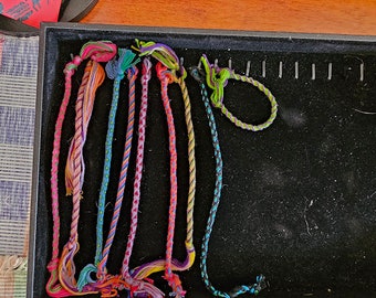 May include: A collection of colorful braided cords, some with tassels, arranged in a tray. The cords are various shades of blue, green, pink, and yellow.