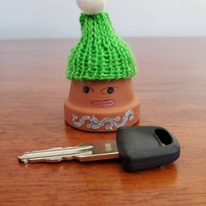 May include: A small terracotta pot with a green knitted hat and a white pom-pom on top. The pot has a painted face with brown eyes, a pink mouth, and a silver glitter design around the rim. A silver key with a black plastic head is lying on a wooden surface.