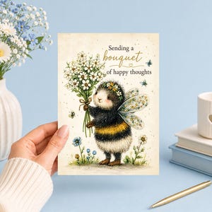 Puede incluir: Una tarjeta de felicitación con la ilustración de un abejorro peludo con alas, que lleva una corona de flores y sostiene un ramo de margaritas blancas. El texto dice, Sending a bouquet of happy thoughts, con pequeñas mariposas azules.