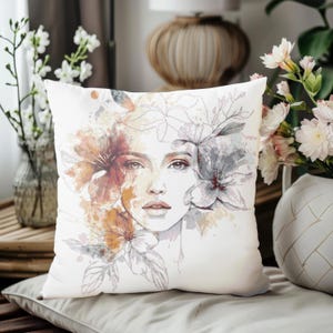 Puede incluir: Cojín cuadrado decorativo con un retrato en acuarela de un rostro femenino rodeado de elementos florales. El diseño incorpora tonos naranja, marrón y gris sobre un fondo blanco. Un artículo de decoración para el hogar.