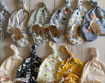 May include: A collection of baby hats and matching mittens in various patterns, including stripes, florals, and animal prints. Each set is tied with a white ribbon and a tag that reads "Babycake Designs."