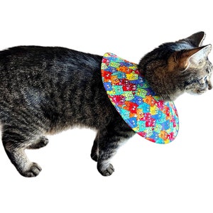 May include: A tabby cat wearing a colorful, multi-patterned, soft, fabric, cone-shaped, protective collar. The collar is covered in a repeating pattern of cartoon cats in various colors.