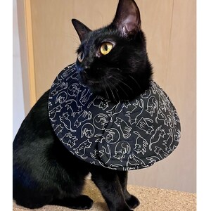 May include: A black cat wearing a black and white patterned Elizabethan collar. The collar features a repeating design of white cat illustrations on a black background. The cat is sitting on a light brown surface.