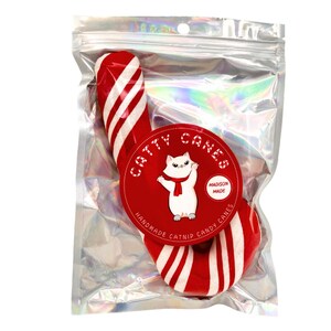 Pode incluir: Um brinquedo de cana de a&ccedil;&uacute;car de catnip vermelho e branco para gatos. A cana de a&ccedil;&uacute;car est&aacute; embalada em um saco de pl&aacute;stico transparente com um r&oacute;tulo que diz "Catty Canes" e "Handmade Catnip Candy Canes".