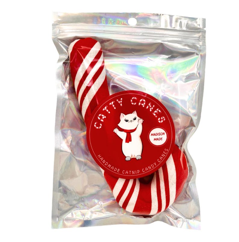 Pode incluir: Um brinquedo de cana de a&ccedil;&uacute;car de catnip vermelho e branco para gatos. A cana de a&ccedil;&uacute;car est&aacute; embalada em um saco de pl&aacute;stico transparente com um r&oacute;tulo que diz "Catty Canes" e "Handmade Catnip Candy Canes".