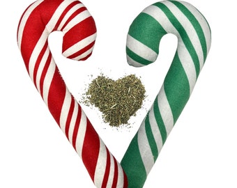 May include: Two candy cane-shaped cat toys, one red and white striped, the other green and white striped, arranged to form a heart shape. A pile of dried catnip is in the center. The toys are made of fabric and are designed for feline play.