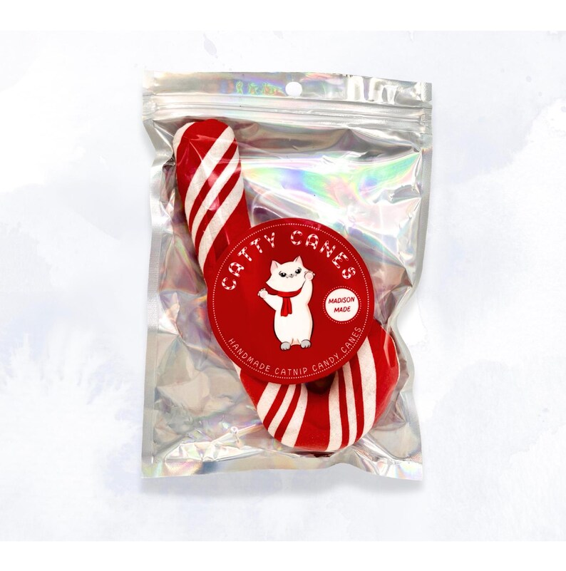 Pode incluir: Um brinquedo de erva-dos-gatos em forma de bengala de caramelo vermelho e branco com um gato branco usando uma echarpe vermelha em um c&iacute;rculo vermelho com o texto "Catty Canes" e "Madison Made Candy Canes".