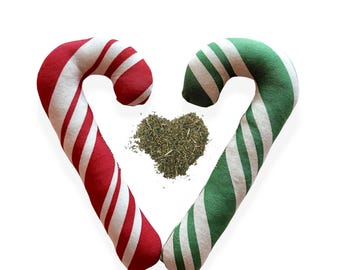 May include: Two fabric candy canes arranged in a heart shape, one with red and white diagonal stripes and the other with green and white diagonal stripes, surrounding a central pile of dried green herbs shaped like a small heart.