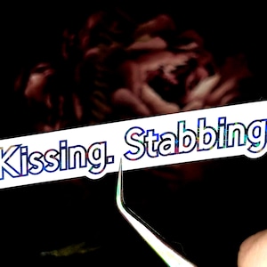 May include: A white sticker with the text "[Kissing. Stabbing.]" in a colorful, holographic font. The sticker is being held by a pair of tweezers against a dark background with a blurred floral design.