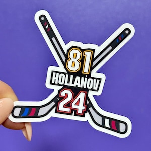 Può includere: Un adesivo con due mazze da hockey incrociate con i numeri 81 e 24. Il nome "HOLLANOV" è stampato sull'adesivo. Le mazze da hockey hanno dettagli neri, bianchi, rossi, blu e grigi. Lo sfondo è viola.
