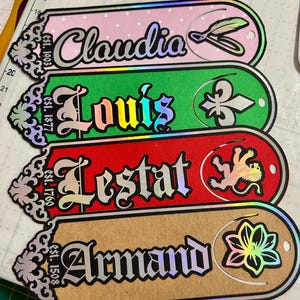 Puede incluir: Cuatro etiquetas decorativas con bordes iridiscentes. Las etiquetas presentan los nombres "Claudia", "Louis", "Lestat" y "Armand" en fuentes ornamentadas. Cada etiqueta tiene un color de fondo único y un emblema decorativo.