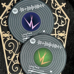 Puede incluir: Dos posavasos redondos con diseño de disco de vinilo. Cada posavasos presenta un círculo central con un logotipo 'W' estilizado y el texto "Long Face The Vampire Lestat". Los posavasos tienen un diseño inspirado en Spotify con una barra de reproducción.