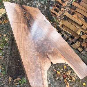 May include: A handcrafted, rectangular wooden table top with a natural edge. The wood grain is a rich brown with darker, swirling patterns. The table top is supported by a tree trunk, with a stack of firewood in the background.