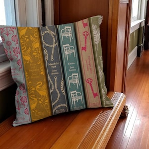 May include: A decorative throw pillow featuring a design of classic book spines. The pillow showcases titles like "Mansfield Park" and "Emma" with decorative elements such as keys, chairs, and floral patterns. The color palette includes shades of blue, yellow, and pink.