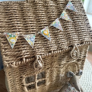 May include: A house-shaped wicker basket with a handle, adorned with a garland of floral pennants. A small fabric mouse in a floral dress stands in front of the basket. The basket is made of woven material.