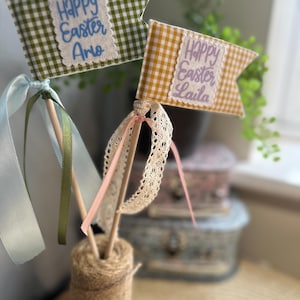 May include: Two Easter flags on wooden sticks. One is green and white gingham with "Happy Easter Arlo" in blue. The other is yellow and white gingham with "Happy Easter Laila" in lavender. Ribbons and lace decorate the sticks.