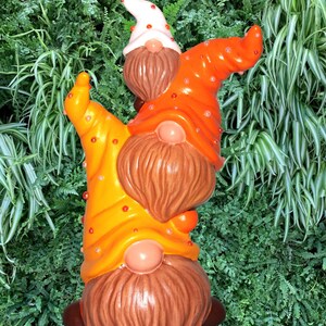 May include: A whimsical garden gnome statue featuring three gnomes with orange, yellow, and white hats adorned with red dots. The gnomes have brown beards and noses. The statue is set against a backdrop of green foliage.