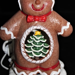 May include: A gingerbread man figurine with white icing and a red bow tie. The figurine has a hole in the center with a green Christmas tree inside.