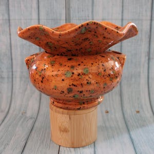 May include: A ceramic planter with a ruffled top edge and a rounded body, featuring a glossy orange glaze with dark brown and green speckles. The planter is supported by a cylindrical light-colored wooden base stand.
