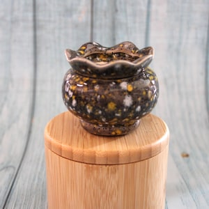 May include: A small, round ceramic jar with a scalloped rim, featuring a dark brown glaze speckled with yellow and white spots. The jar sits atop a cylindrical, light-colored bamboo container with a smooth, vertical wood grain texture.